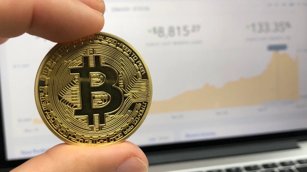 Bitcoin com uma tela ao fundo mostrando histórico de valores da criptomoeda
