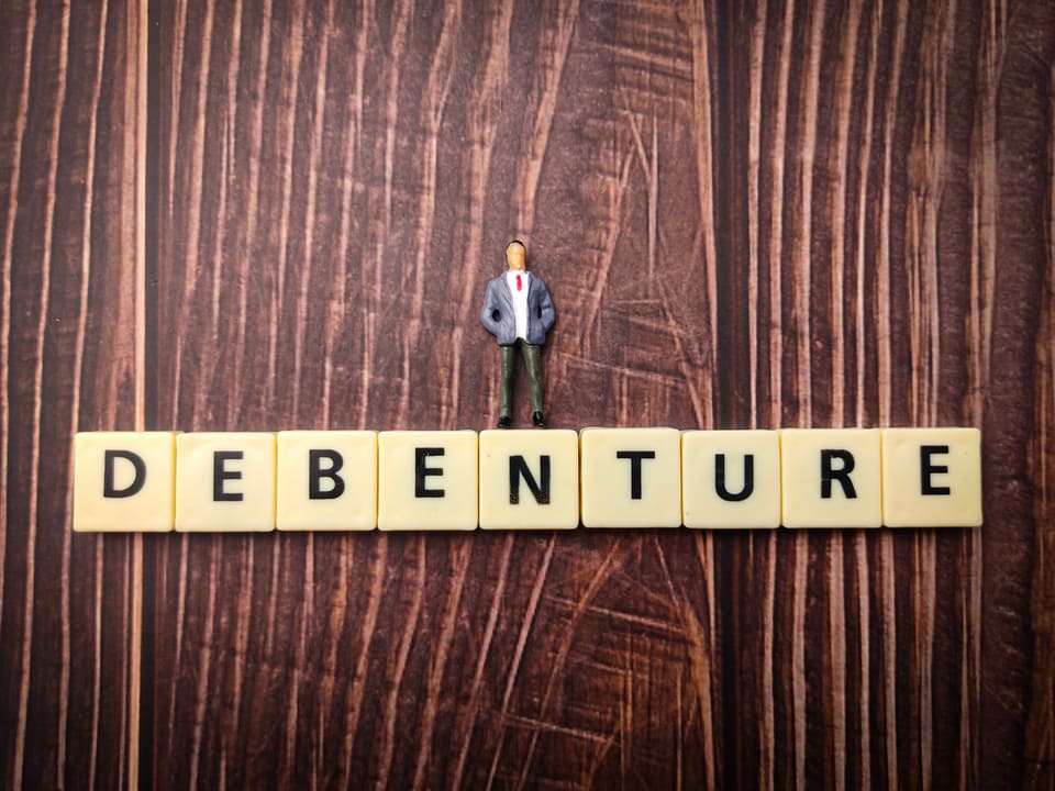 Palavra "debenture" escrita por meio de blocos com letras sob superfície de madeira com boneco de pessoa de terno em cima do termo