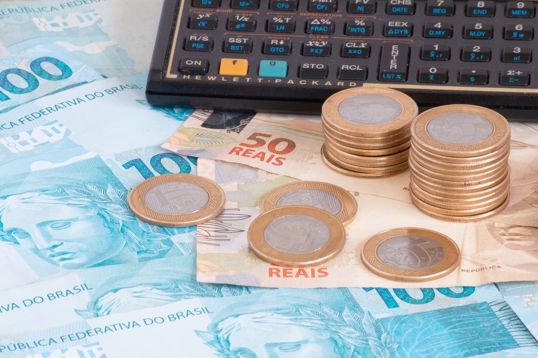Notas de 100 e 50 reais, além de moedas de 1 real, sob superfície junto a calculadora preta