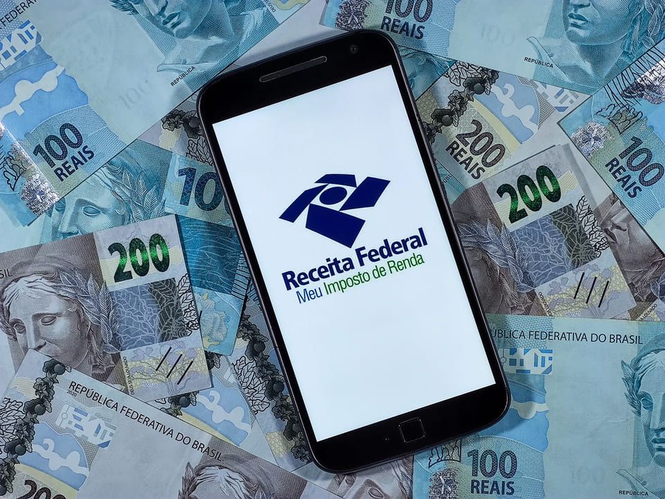 Celular sobre notas de 100 e 200 reais exibe logomarca da Receita Federal 